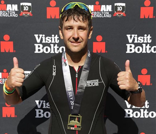 male athlete celebrating with thumbs up after completing an Ironman event wearing a medal and sunglasses in Bolton England endurance sports 2 achievements