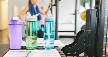 three water bottles in a gym setting with workout equipment and a person in the background hydrating after exercise seven hydration tips